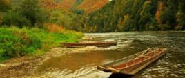 Na pierwszym planie górska rzeka z zacumowanymi łodziami, w tle zalesione góry w jesiennej szacie.