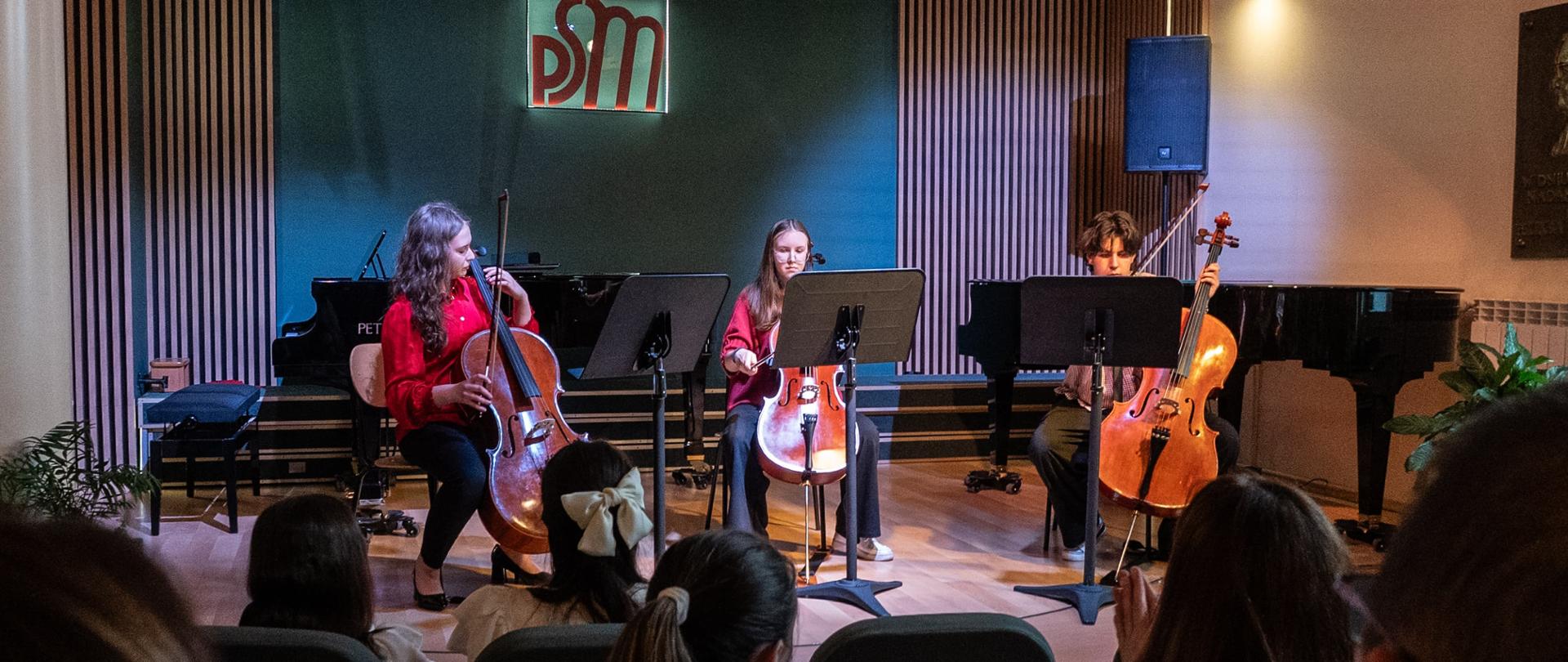 Na zdjęciu widać troje młodych muzyków siedzących na scenie i grających na wiolonczelach. Każdy z nich ma przed sobą pulpit z nutami. W tle widnieje logo szkoły muzycznej „PSM” zawieszone na ciemnozielonej ścianie. Po bokach sceny znajdują się fortepiany. Przed sceną siedzi publiczność, która z uwagą słucha występu, a niektórzy widzowie biją brawo. Atmosfera jest skupiona i pełna uznania, charakterystyczna dla koncertów szkolnych i kameralnych występów muzycznych.