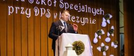 Minister Czarnek stoi przy mównicy i mówi do mikrofonu, za nim złota kotara a na niej niebieski napis 25-lecie Zespołu Szkól specjalnych przy DPS w Matczynie.