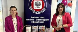Zdjęcie przedstawia stoisko edukacyjne Powiatowej Stacji Sanitarno-Epidemiologicznej w Siemiatyczach podczas wydarzenia profilaktycznego. Na stole znajdują się materiały informacyjne dotyczące profilaktyki nowotworów, szczepień przeciw HPV oraz modele do nauki samobadania piersi. Za stołem stoją dwie pracownice PSSE.