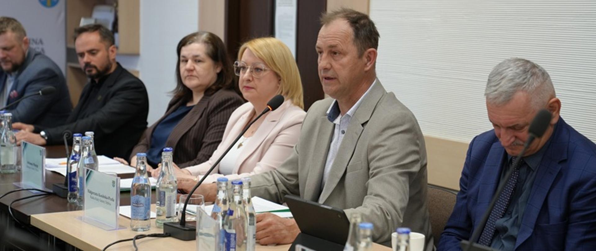 Grupa pięciu osób siedzi przy długim stole konferencyjnym w jasnym pomieszczeniu. Na pierwszym planie po prawej stronie mężczyzna w jasnej marynarce mówi do mikrofonu, patrząc przed siebie. Przed nim stoją szklane butelki z wodą, tablet oraz kartka z nazwiskiem. Obok niego siedzi kobieta w jasnoróżowej marynarce i okularach, zwrócona w stronę mówcy. Dalej widoczna jest kobieta w ciemnej marynarce oraz dwóch mężczyzn w garniturach. Na stole ustawiono mikrofony, szklanki i butelki wody dla każdego uczestnika. W tle widać półki, drzwi oraz fragment ściany z oznaczeniami instytucji.