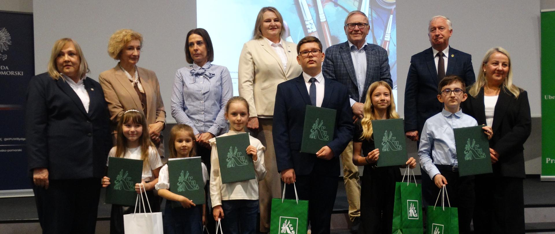 Szóstka dzieci pozuje do zdjęcia. Dzieci trzymają w rękach dyplomy z logo KRUS oraz torby prezentowe. Dzieci są laureatami XV Ogólnopolskiego Konkursu Plastycznego dla Dzieci WIOSKI BEZ TROSKI. Za dziećmi stoją przedstawiciele instytucji organizujących konkurs.