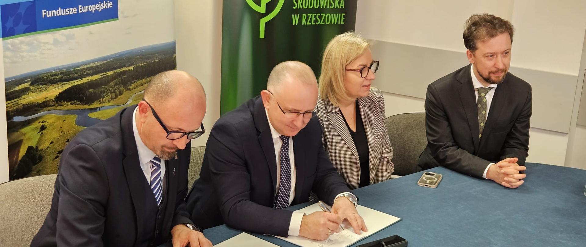 Oficjalny moment podpisania dokumentów. Dwóch mężczyzn siedzi przy stole i składa podpisy, podczas gdy kobieta w okularach oraz mężczyzna z brodą, którzy również siedzą przy stole przyglądają się temu procesowi. 