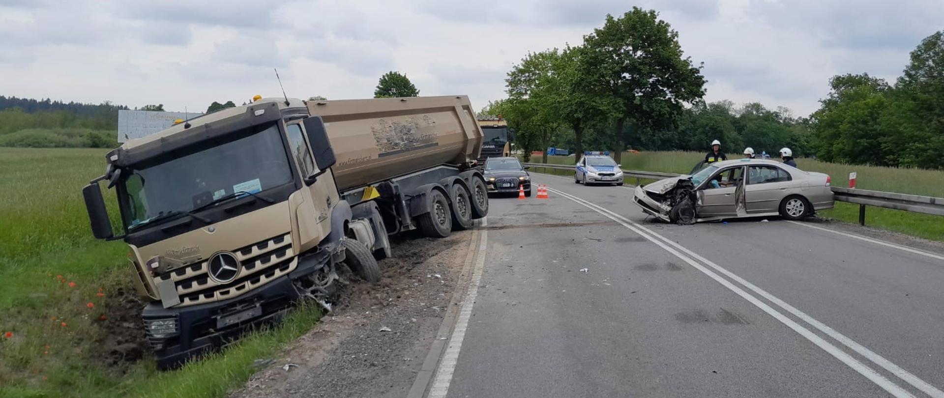 Miejsce wypadku. Z lewej strony na poboczu drogi znajduje się uszkodzony ciągnik siodłowy z naczepą. Z prawej strony na jezdni znajduje się uszkodzony samochód osobowy.