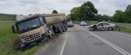 Miejsce wypadku. Z lewej strony na poboczu drogi znajduje się uszkodzony ciągnik siodłowy z naczepą. Z prawej strony na jezdni znajduje się uszkodzony samochód osobowy.