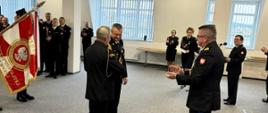 Strażak w mundurze wyjściowym stoi przed dwoma funkcjonariuszami Państwowej Straży Pożarnej w mundurach wyjściowych za nimi jest sztandar obok stoi kadra kierownicza Państwowej Straży Pożarnej województwa pomorskiego.