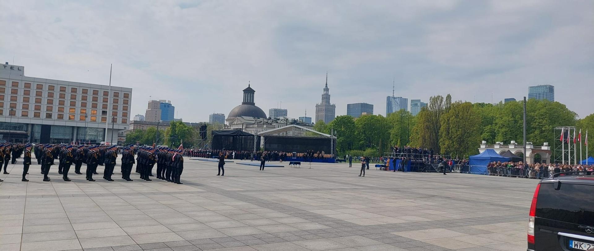 Zdjęcie przedstawia plac Marszałka Józefa Piłsudskiego w Warszawie. W kolumnie stoją strażacy w galowych mundurach. Przed nimi stoi pusta scena, dookoła widać tłum widzów.