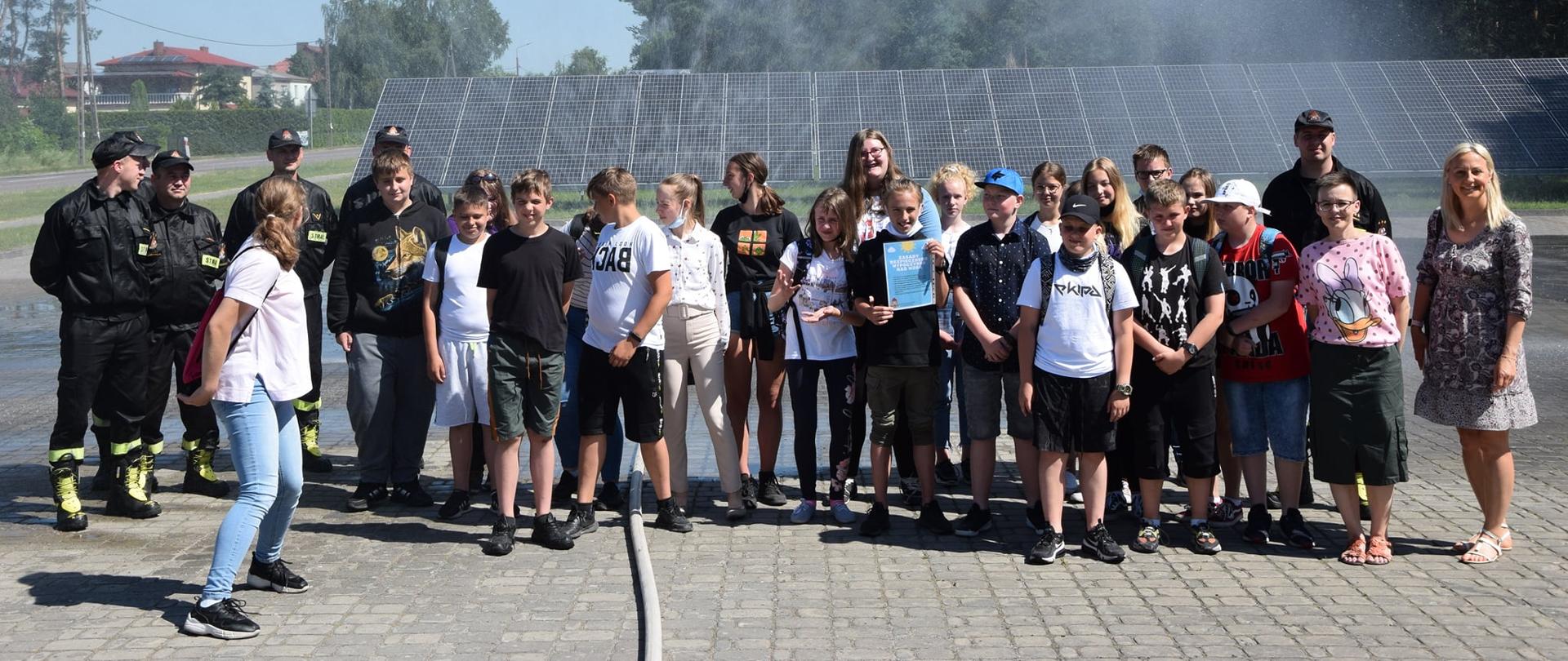 JEST TO ZDJĘCIE GRUPOWE, NA KÓRYM OD LEWEJ STRONYSTRAŻAKÓW W UBRANIACH KOSZAROWYCH, NASTĘPNIE DZIECI A ZA NIMI DWIE DOROSŁE KOBIETY. W TLE WIDAC NAZIEMNĄ INSTALACJĘ FOTOWOLTAICZNĄ, LAS I FRAGMENTY BUDYNKÓW.