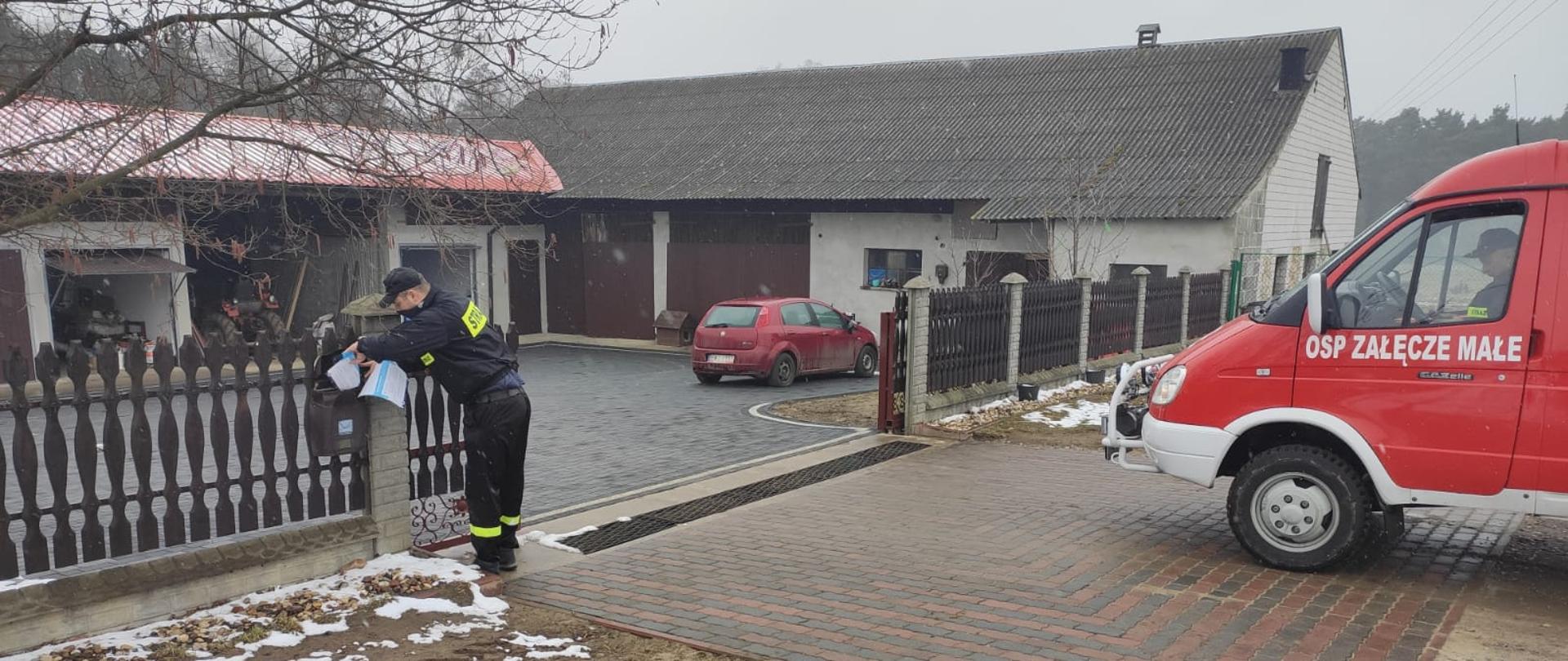 Zdjęcie przedstawia strażaka w ubraniu koszarowym wkładającego ulotkę do skrzynki na listy, widoczny jest także samochód pożarniczy