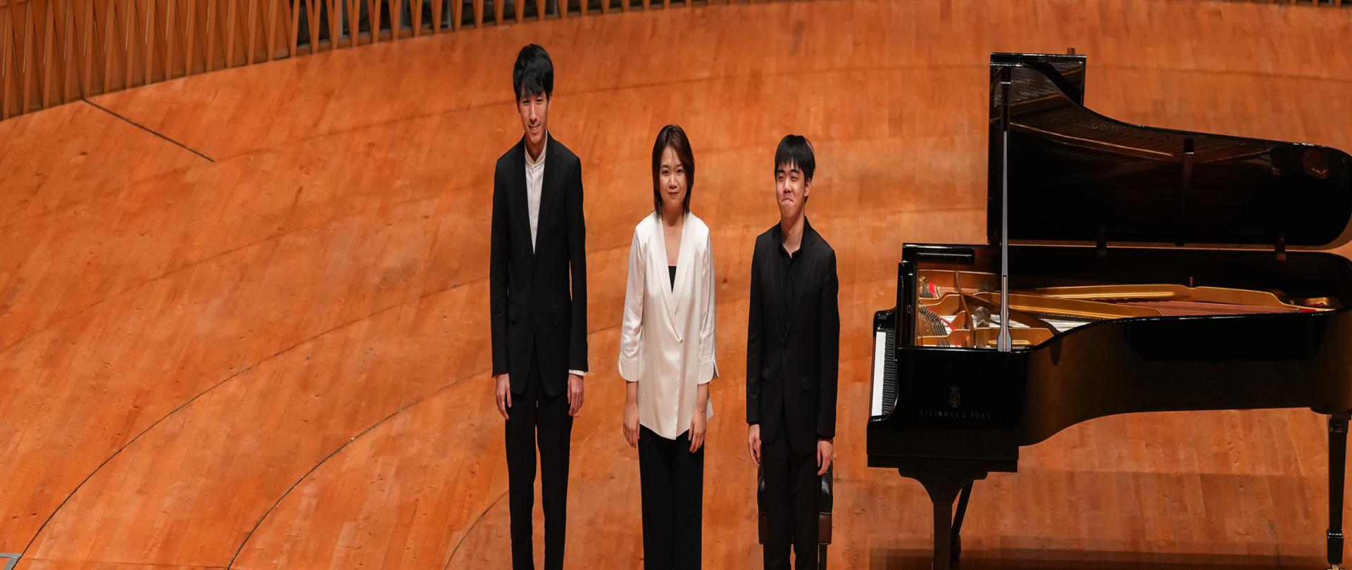 trzech pianistów stoi na scenie obok fortepianu