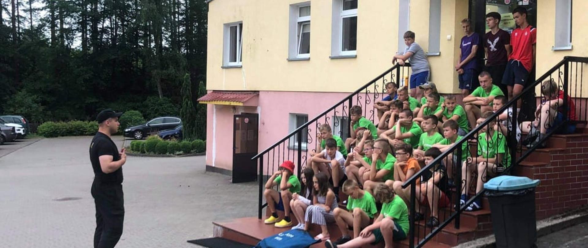 Zdjęcie przedstawia młodzież siedzącą na schodach przed budynkiem szkoły oraz strażaka który udziela instruktażu na temat bezpiecznych wakacji. 