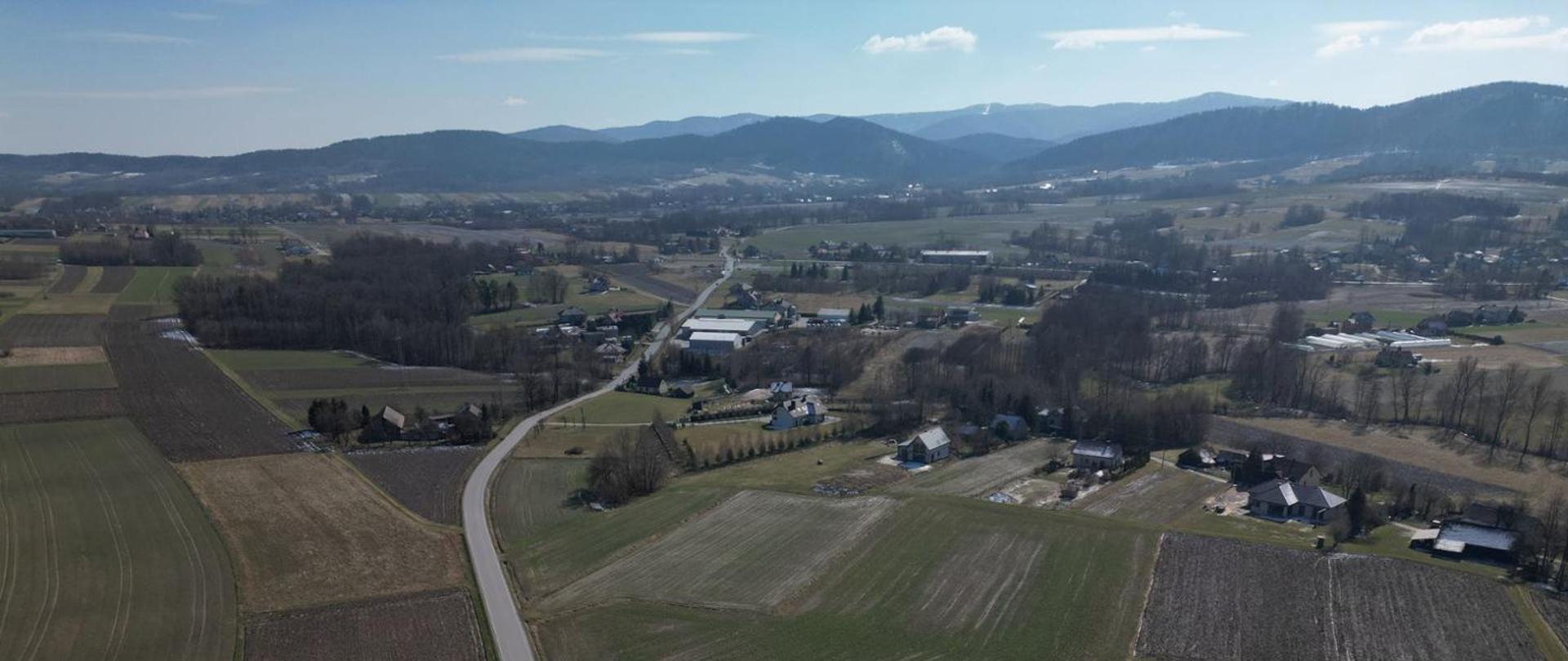 Widok z lotu ptaka na drogę przebiegającą przez niewielką miejscowość. W tle góry.