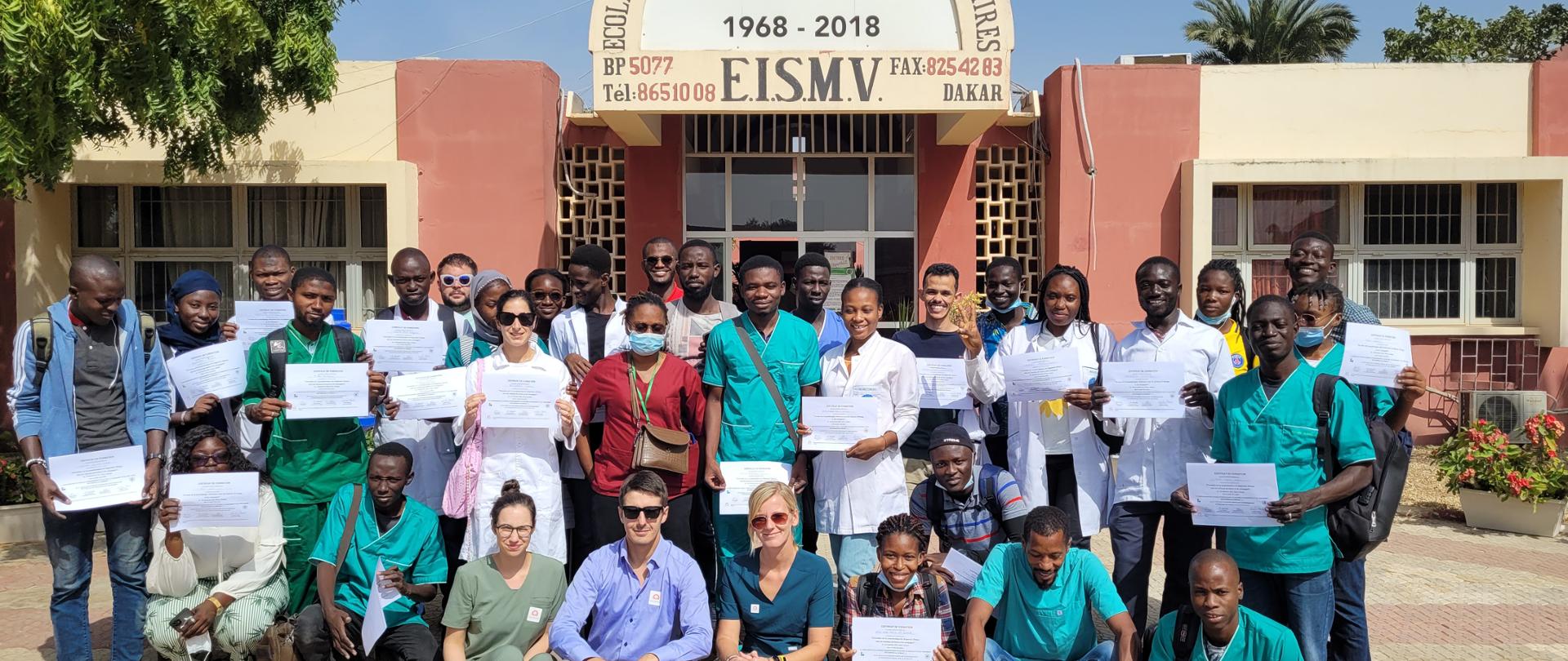 Grupa kobiet i mężczyzn w kitlach pozuje z dyplomami przed budynkiem szpitala.