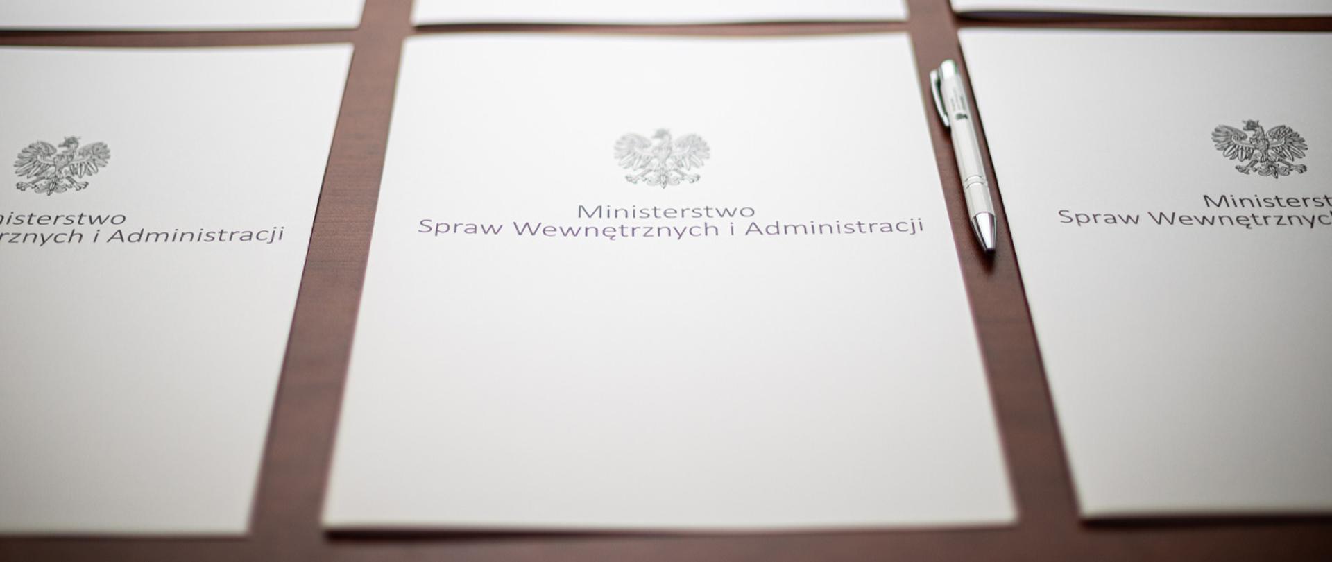  Cały kadr wypełniają ułożone obok siebie teczki z napisem "Ministerstwo Spraw Wewnętrznych i i Administracji" i orłem z godła narodowego. Obok jednej z teczek leży długopis MSWiA
