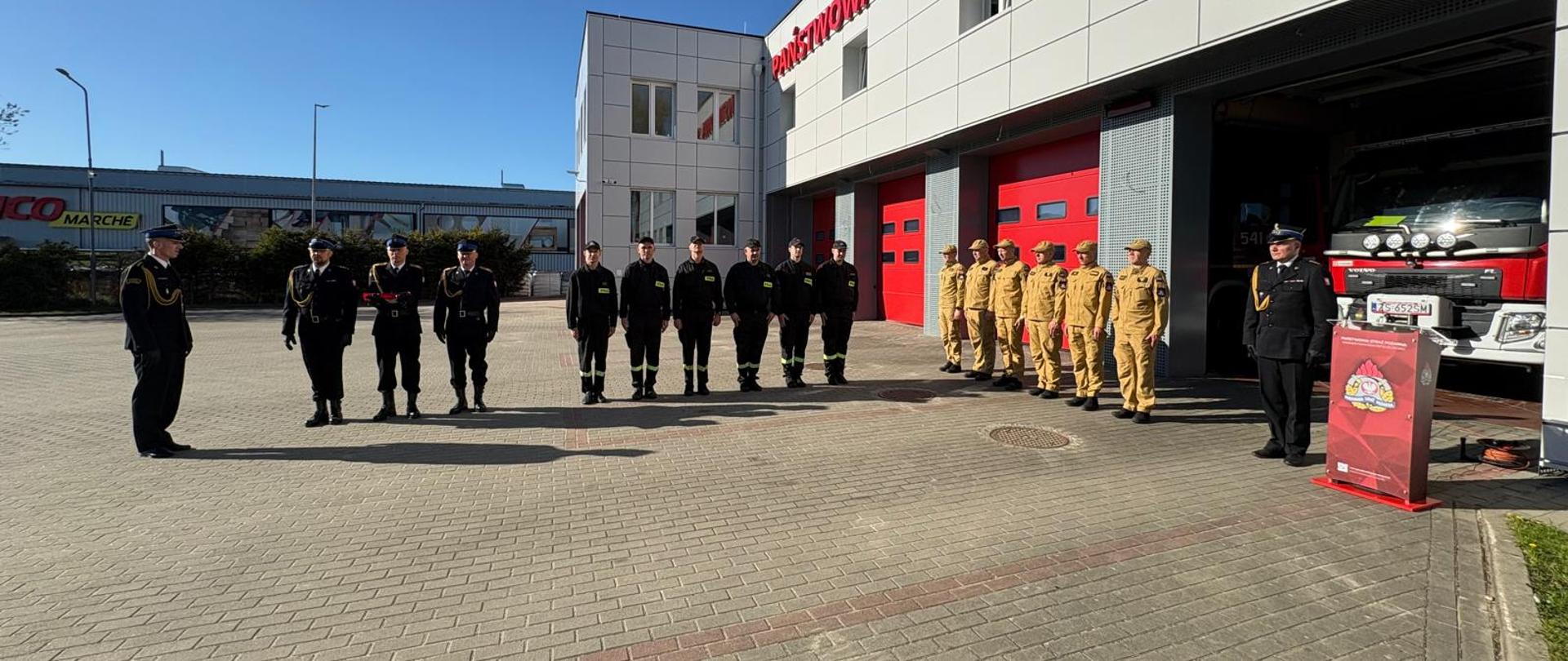 Uroczyste podniesienie flagi państwowej przed budynkiem straży pożarnej. Na placu wyłożonym kostką brukową stoją w równych szeregach strażacy w galowych i służbowych mundurach – część w czarnych, część w piaskowych. Po prawej stronie widoczny jest czerwony wóz strażacki zaparkowany w otwartym garażu oraz podium z mikrofonem. Na tle nowoczesnego budynku strażnicy, oznaczonego czerwonym napisem, uczestnicy ceremonii zachowują powagę i skupienie w słoneczny, bezchmurny dzień.