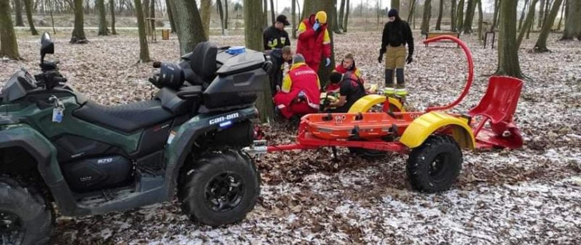 Centralny punkt zdjęcia stanowi quad i przyczepka do transportu osoby poszkodowanej. W tle za zestawem pojazdów stoją ratownicy WOPR oraz strażak. Wszystkie osoby pomagają osobie poszukiwanej leżącej na przyczepce