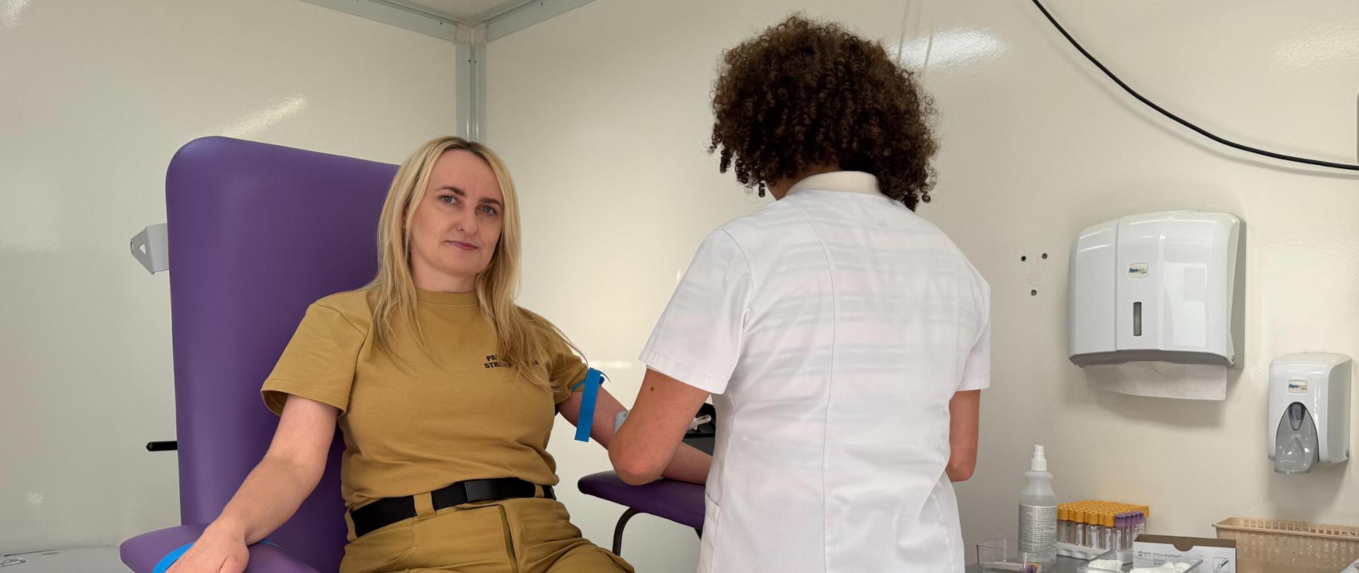 Blondynka w piaskowej koszulce i spodniach mundurowych siedzi na fioletowym fotelu zabiegowym i patrzy w stronę obiektywu. Na jej lewym ramieniu założona jest niebieska opaska uciskowa (staza). Przed nią stoi tyłem pracownica medyczna w białym kitlu i z kręconymi włosami, wykonująca zabieg pobrania krwi. Akcja dzieje się w jasnym, mobilnym punkcie medycznym, w którym widoczne są białe meble, pojemniki z probówkami, rękawiczki jednorazowe oraz sprzęt medyczny umieszczony na blacie.