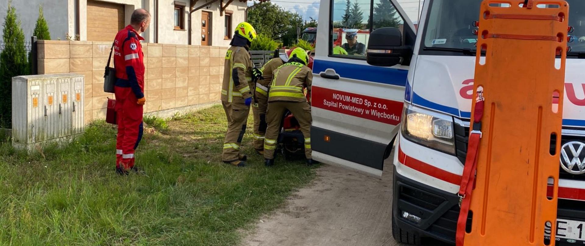 Zdjęcie wykonano porą dzienną, wykonano przy ulicy Wojska Polskiego w Więcborku. Na zdjęciu widać tyłem 3 strażaków udzielających pomocy poszkodowanemu po wypadku. Bokiem stoi ratownik medyczny , natomiast przodem stoi policjant. Po prawej stronie kartka pogotowia ratunkowego o którą oparte są nosze typu deska. W tle budynek mieszkalny. 