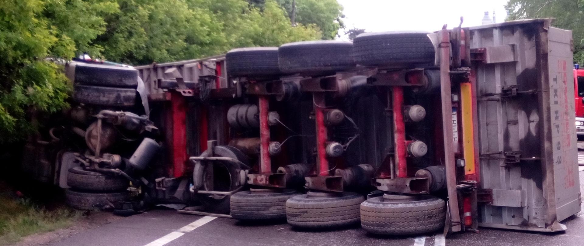 Zdjęcie przedstawia przewrócony ciągnik siodłowy oraz naczepę na drodze. W tle widać przydrożne drzewa.