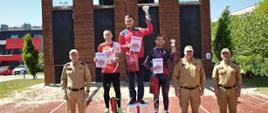 W rzędzie przed brązowo - czarno wspinalnią stoją strażacy ubrani w piaskowe mundury oraz sportowcy ubrani w dresy. Sportowcy stoją na podwyższeniach w rękach trzymają dyplomy i puchary.