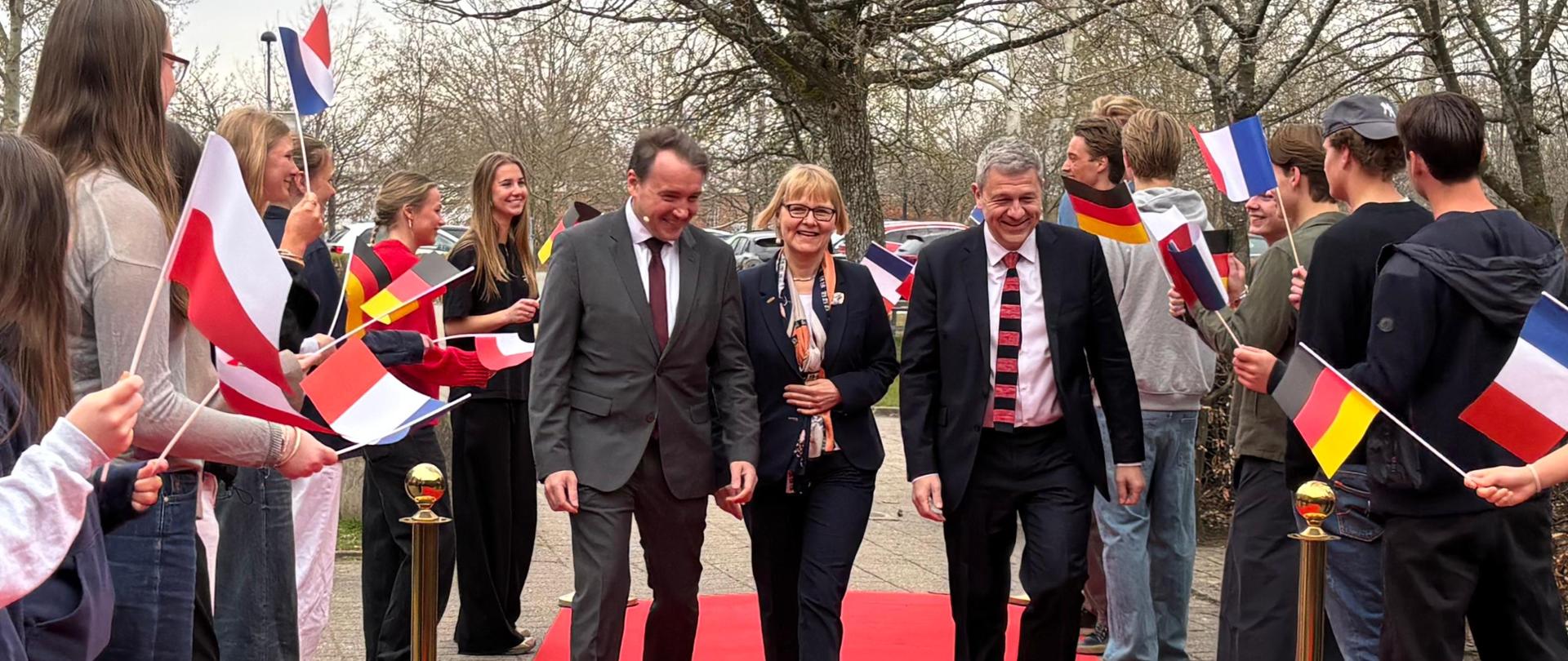 Grupa osób przechodzi po czerwonym dywanie, a po obu stronach stoją młodzi ludzie trzymający flagi.