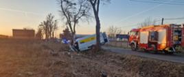 Zdjęcie przedstawia:
od lewej strony biały samochód typu bus po uderzeniu w drzewo, następnie czerwony wóz straży pożarnej