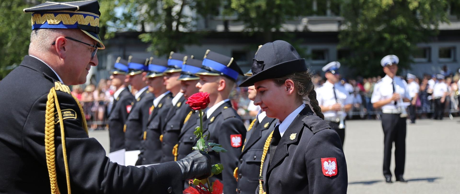 Po lewo z-ca komendanta głównego PSP , po prawo naprzeciw stoją w szeregu strażacy do promocji . Jako pierwsza stoi kobieta , której z-ca KG PSP podaje na zdjęciu różę