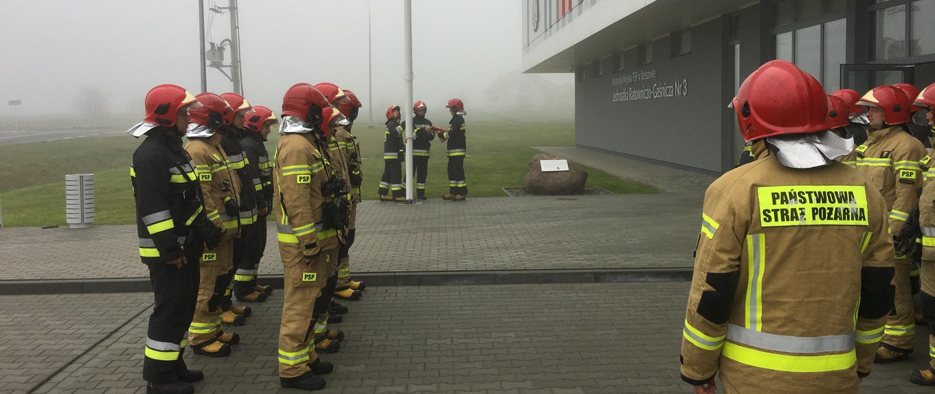 Zdjęcie przedstawia strażaków w umundurowaniu bojowym zmiany zdającej i przejmującej Jednostki Ratowniczo-Gaśniczej Nr 3 w Rzeszowie, stojących naprzeciwko siebie przed budynkiem strażnicy. Trzech z nich podnosi flagę na maszt. 