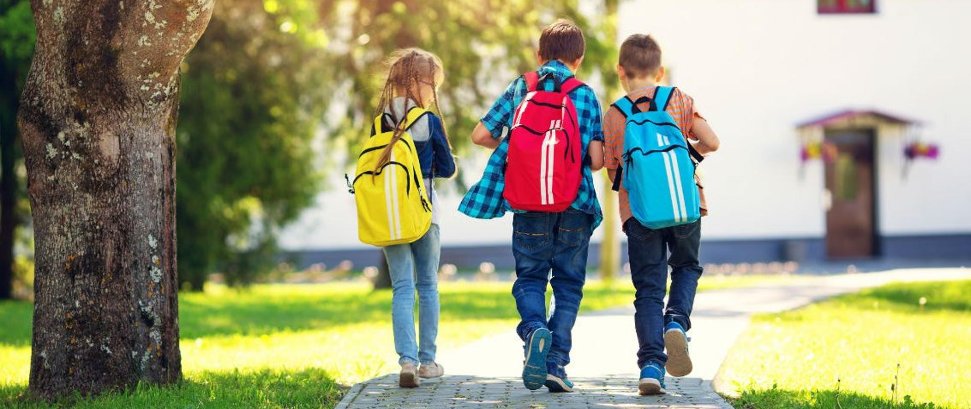 Na zdjęciu znajduje się trójka dzieci, która zmierza w kierunku szkoły, ich atrybutem są tornistry. Dziewczynka wraz z dwoma chłopcami znajdują się na chodniku w parku miejskim. W tle widoczny jest budynek.