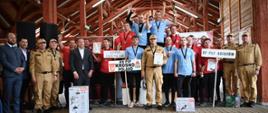 Wspólne zdjęcie grupowe na zakończenie zawodów pożarniczych. Na podium stoją drużyny z KM PSP Krosno (na najwyższym stopniu), KP PSP Jarosław oraz KP PSP Brzozów. Uczestnicy noszą medale i trzymają puchary, dyplomy oraz nagrody rzeczowe. W tle i po bokach stoją funkcjonariusze PSP oraz przedstawiciele organizatorów w mundurach i garniturach. Całość odbywa się w drewnianej hali.