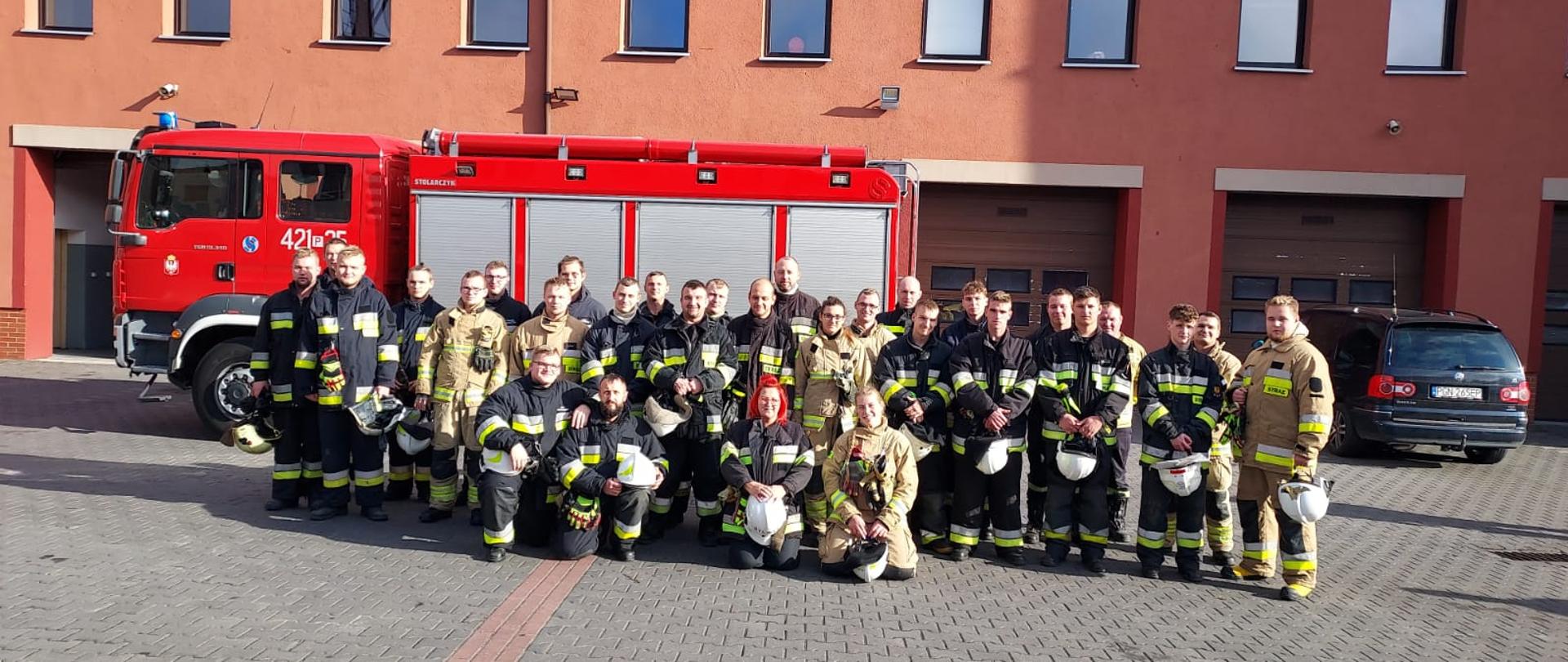Zdjęcie grupowe osób biorących udział w szkoleniu. Druhowie ubrani są w ubrania specjalne typu Nomex i stoją na tle samochodu gaśniczego