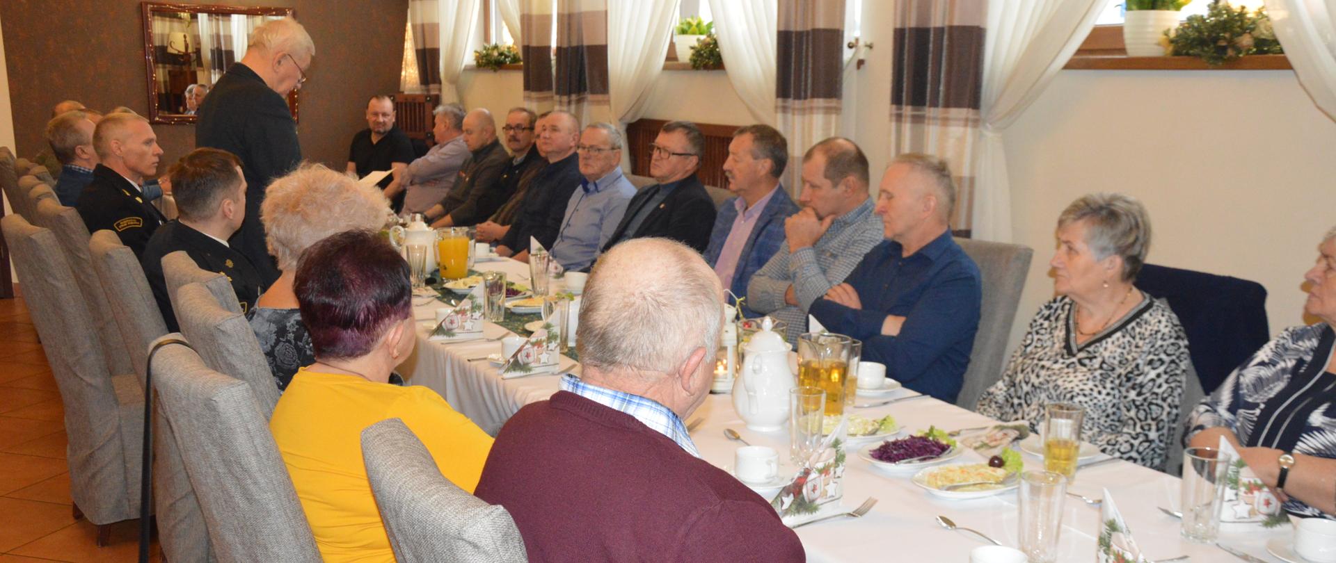 Po obu stronach stołów siedzą uczestnicy spotkania - kobiety i mężczyźni. Wśród nich jest dwóch strażaków PSP, w mundurach wyjściowych. W pozycji stojącej przemawia prezes koła emerytów. Stoły są nakryte białymi obrusami. Stoją na nich filiżanki, szklanki, dzbanki oraz leżą talerzyki z sałatkami. 