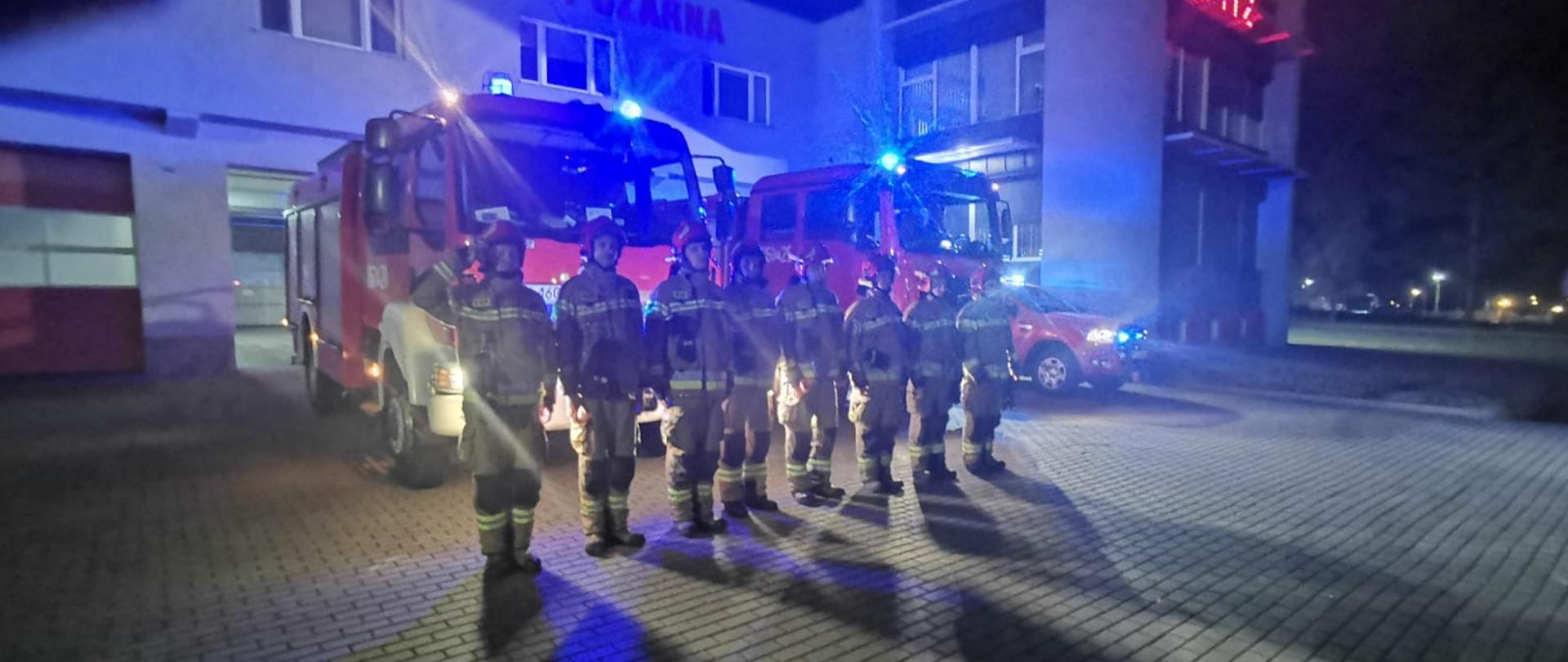 Na zdjęciu widać strażaków, którzy stoją przed jednostką. Samochody strażackie mają włączone sygnały świetlne i dźwiękowe. Strażacy uczcili pamięć zmarłych strażaków. 