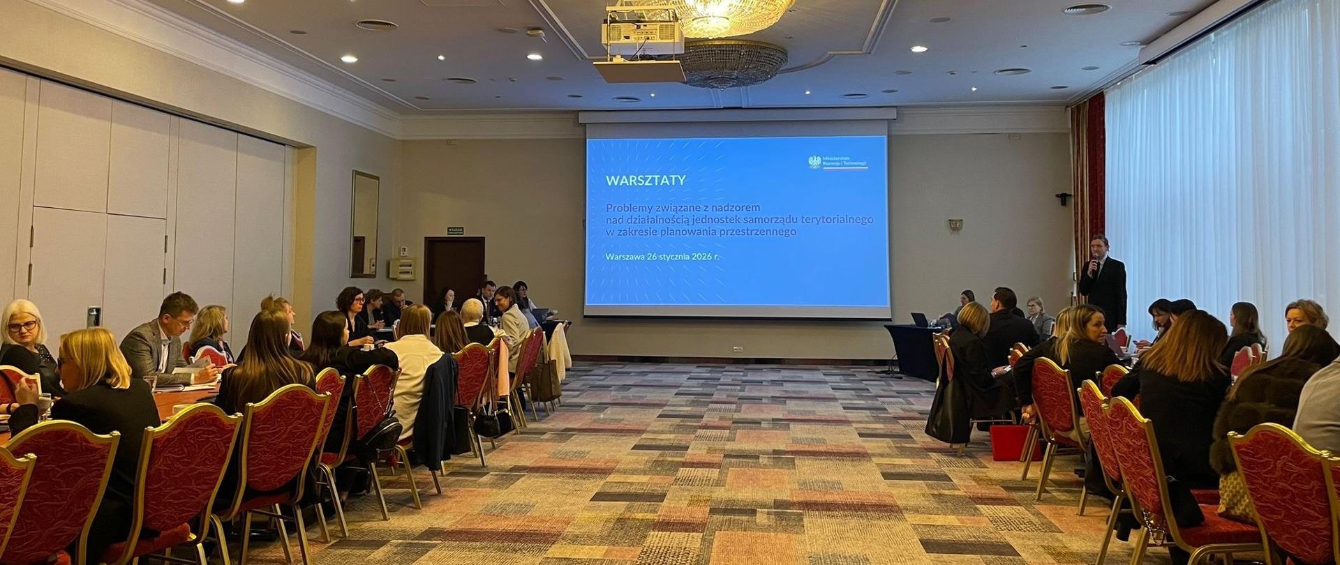 Fotografia przedstawia salę konferencyjną. Po lewej i po prawej stronie siedzą uczestnicy spotkania na krzesłach przy stołach konferencyjnych. W oddali widać wyświetlaną prezentację na projektorze.
