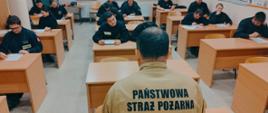 Widok z przodu. Sala egzaminacyjna. Egzaminowani pochylają się nad testami. Nad przebiegiem egzaminu czuwa przewodniczący komisji. 