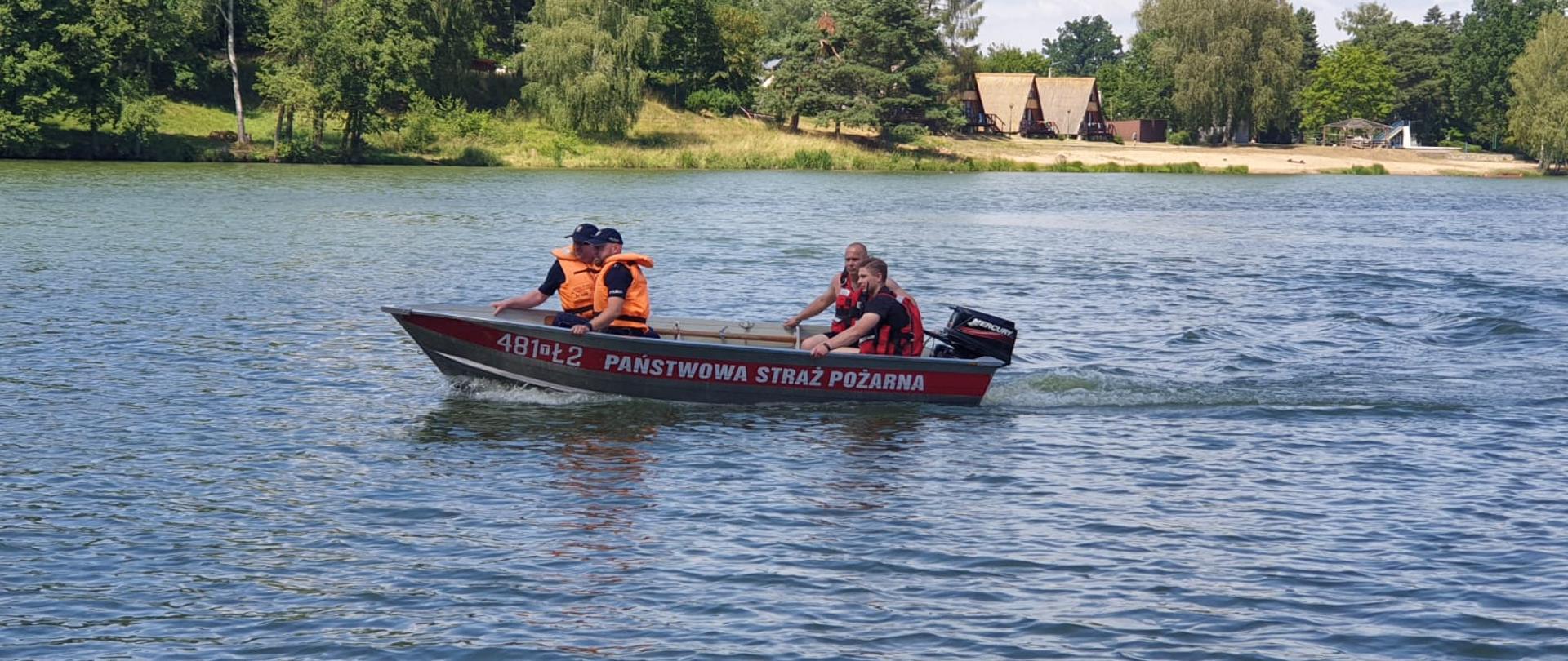 Zdjęcie obrazuje strażaków ćwiczących podejmowanie z wody osoby poszkodowanej. Strażacy ćwiczą podpłynięcie do koła ratunkowego unoszącego się na wodzie i jego podjęcie z wody. 