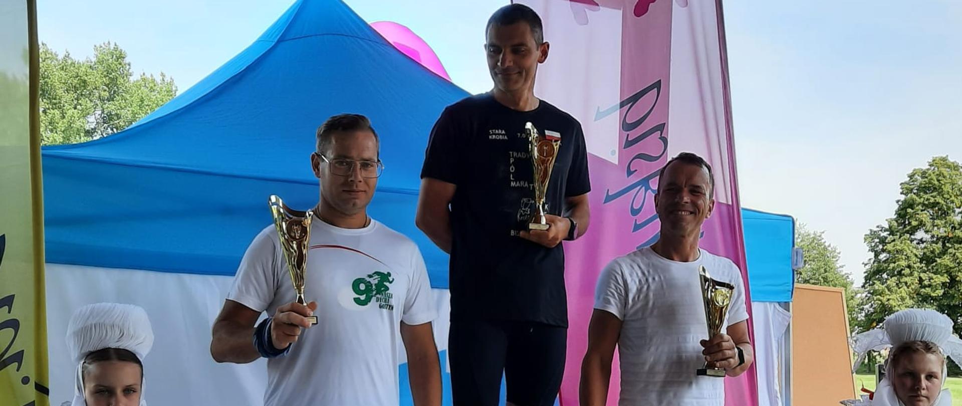Trzech mężczyzn stoi na podium po zakończonym biegu. Każdy z nim trzyma puchar odpowiedni do zajętego miejsca. Obok nich z prawej i lewej strony stoją dwie kobiety ubrane w stroje ludowe.