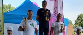 Trzech mężczyzn stoi na podium po zakończonym biegu. Każdy z nim trzyma puchar odpowiedni do zajętego miejsca. Obok nich z prawej i lewej strony stoją dwie kobiety ubrane w stroje ludowe.