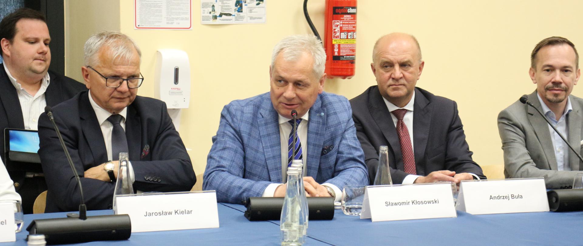 Wojewoda opolski podczas posiedzenia senackiej komisji w Kluczborku. Wojewoda siedzi przy stole w towarzystwie pozostałych uczestników spotkania. Przed wojewodą na stole leży mikrofon. W tle okno oraz gaśnica, ulokowana na ścianie.