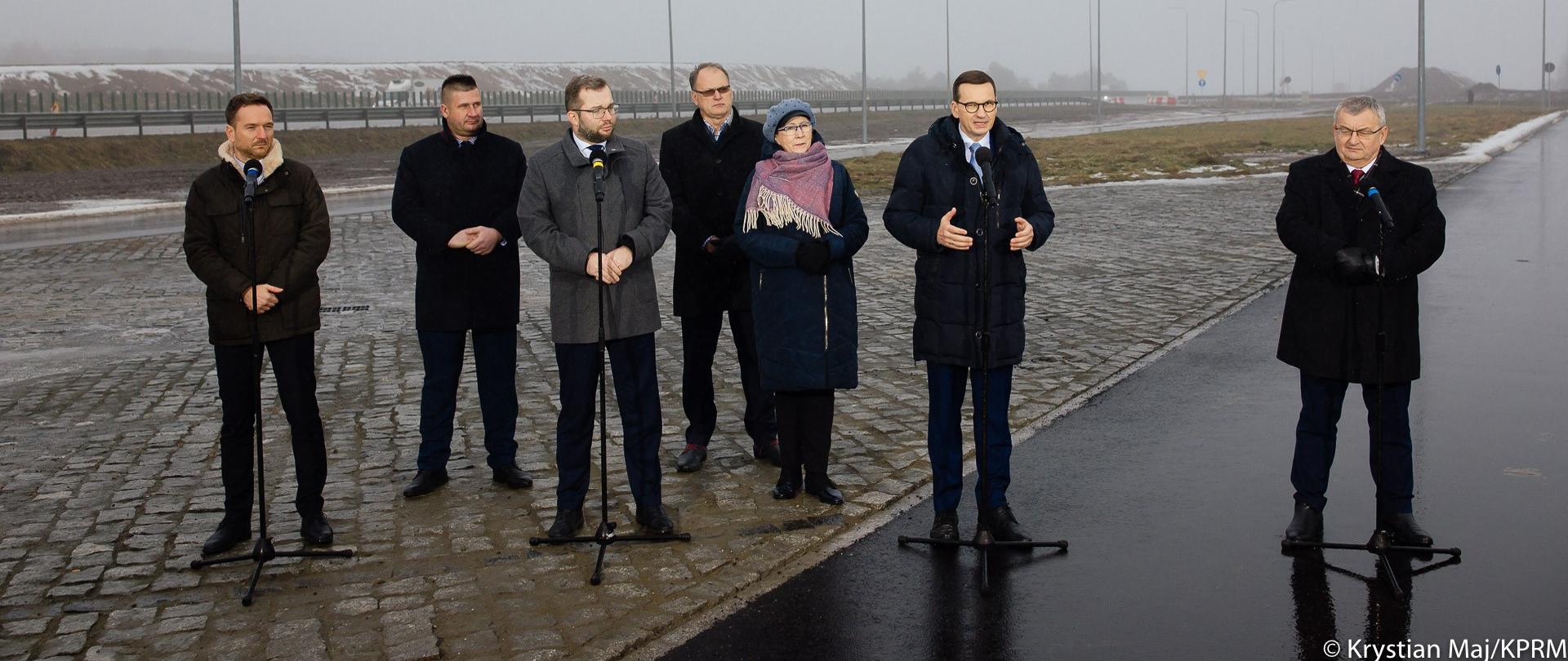Waldemar Buda, Grzegorz Puda, premier Mateusz Morawiecki i Andrzej Adamczyk stoją przy mikrofonach na tle drogi.