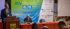 Wiceminister Murdzek stoi za drewnianą mównicą i mówi do mikrofonu, obok mównicy przykryty czerwonym materiałem stół za którym siedzi mężczyzna w garniturze i okularach, za nimi rozwijany baner z napisem XIV konferencja naukowa technologia elektronowa ELTE 2023, przed nimi widać siedzących na ustawionych w rzędy krzesłach ludzi.