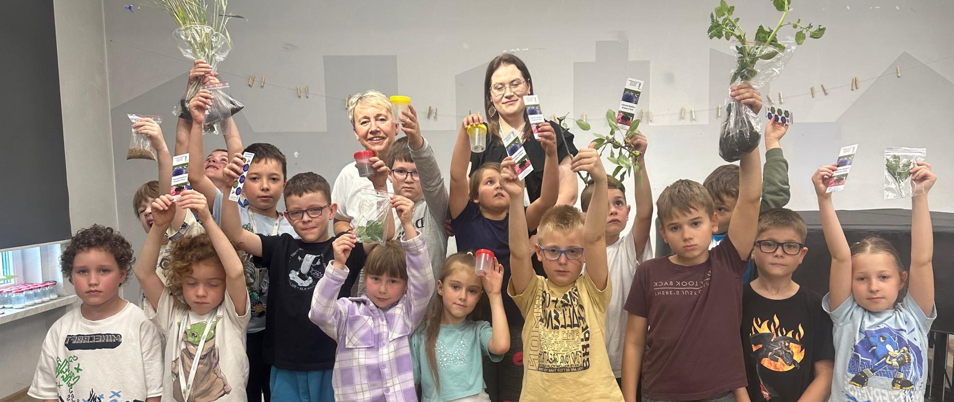 zbiorowe zdjęcie uczniów. Niektóre z dzieci trzymają w uniesionych dłoniach fragmenty roślin, nasiona w woreczkach oraz pojemniki z okazami owadów