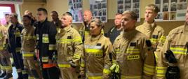 Grupa strażaków w ubraniach bojowych stoi w szeregu w sali remizy podczas odprawy lub szkolenia. Na ścianach za nimi wiszą plansze ze zdjęciami z działań strażackich.