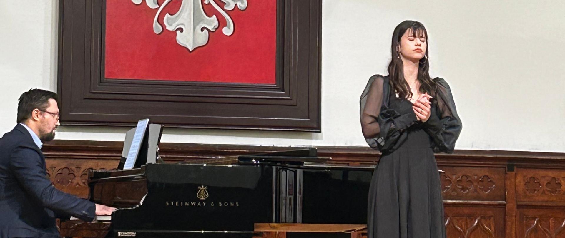 Uczennica w czarnej sukni śpiewa na scenie Sali Królewskiej podczas recitalu dyplomowego, a nauczyciel akompaniuje na fortepianie Steinway, w tle herb Polski.