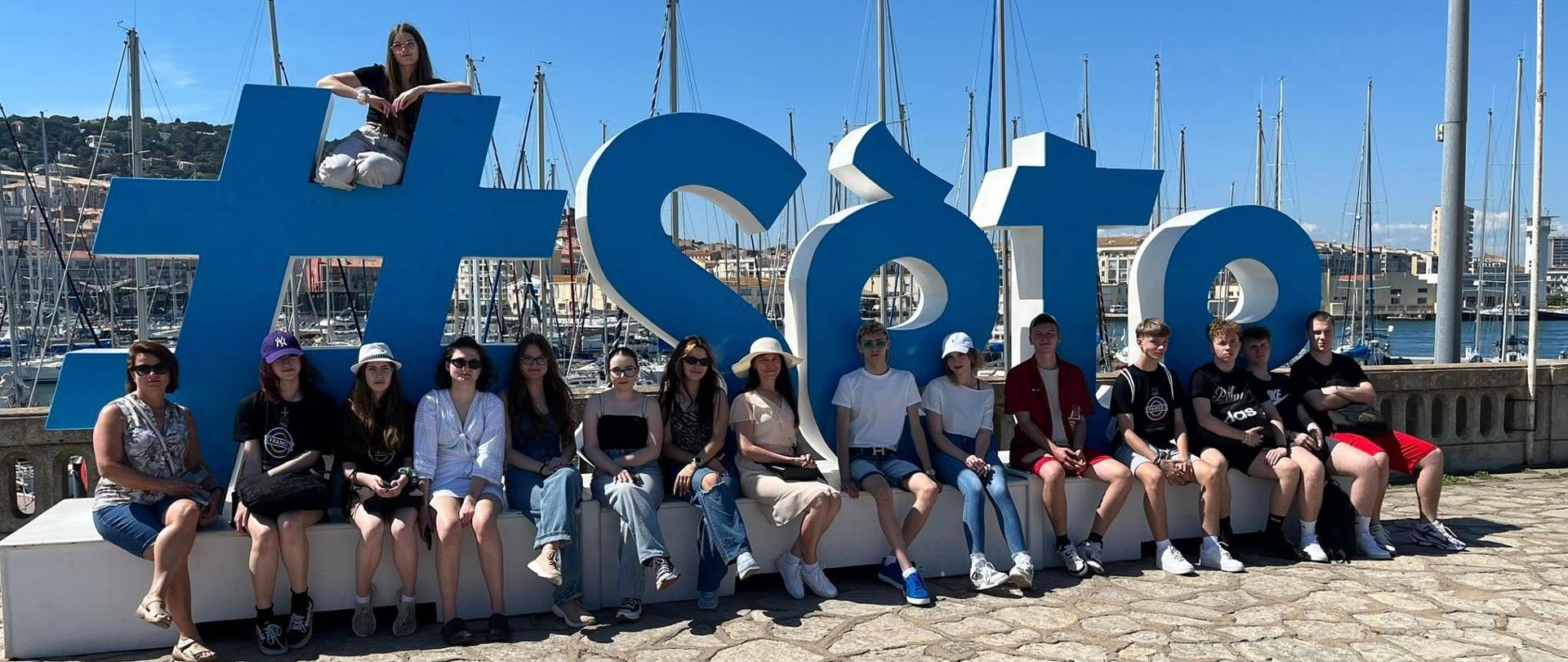 Grupa młodzieży siedzi przy dużym napisie miasta Sète w porcie jachtowym.