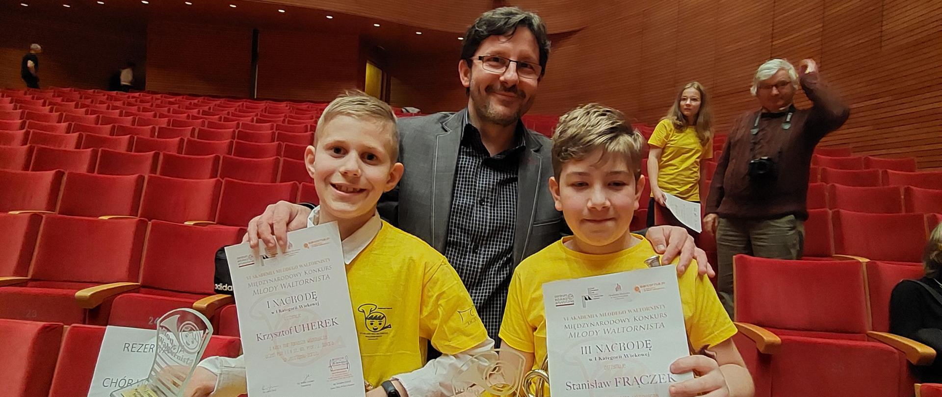 Dwóch chłopców w żółtych koszulkach trzyma waltornie, statuetki i dyplomy, za nimi stoi nauczyciel.
