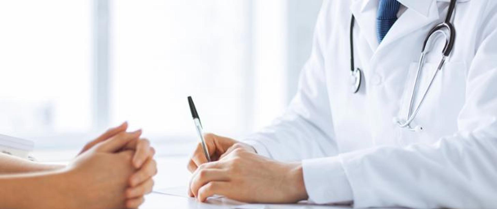 Lekarz w białym fartuchu udziela porady pacjentowi, siedząc naprzeciwko niego przy biurku.