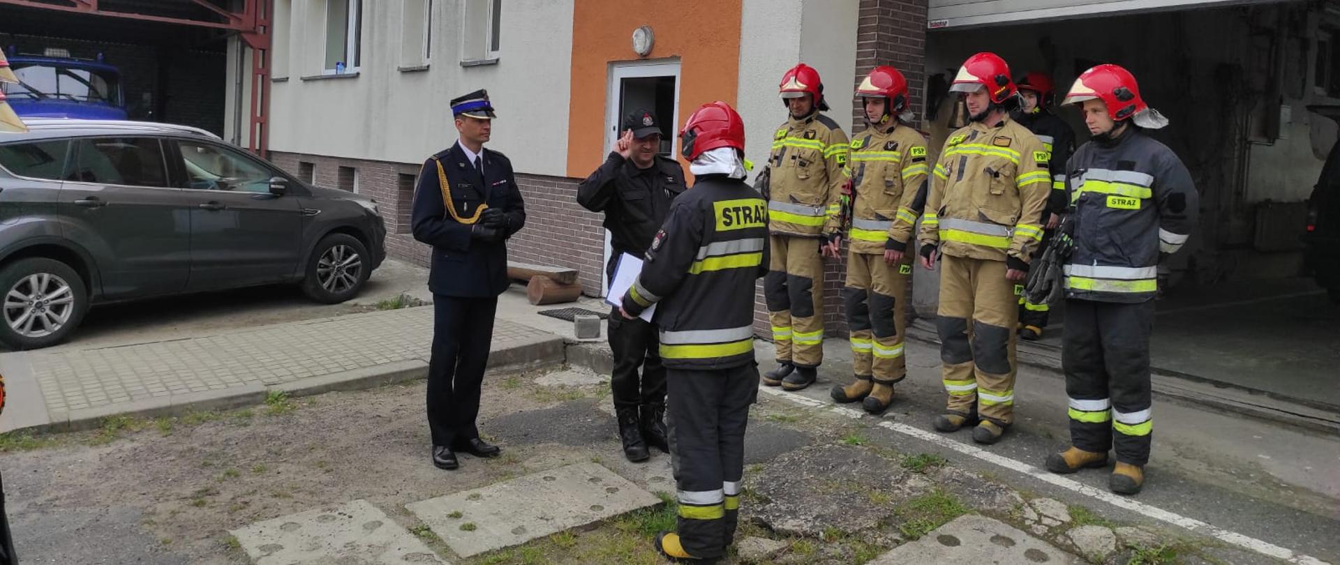 Strażak w umundurowaniu bojowym otrzymuje od swoich przełożonych ubranych w umundurowanie wyjściowe oraz sztabowe list pochwalny. W tle stoją w szeregu inni 