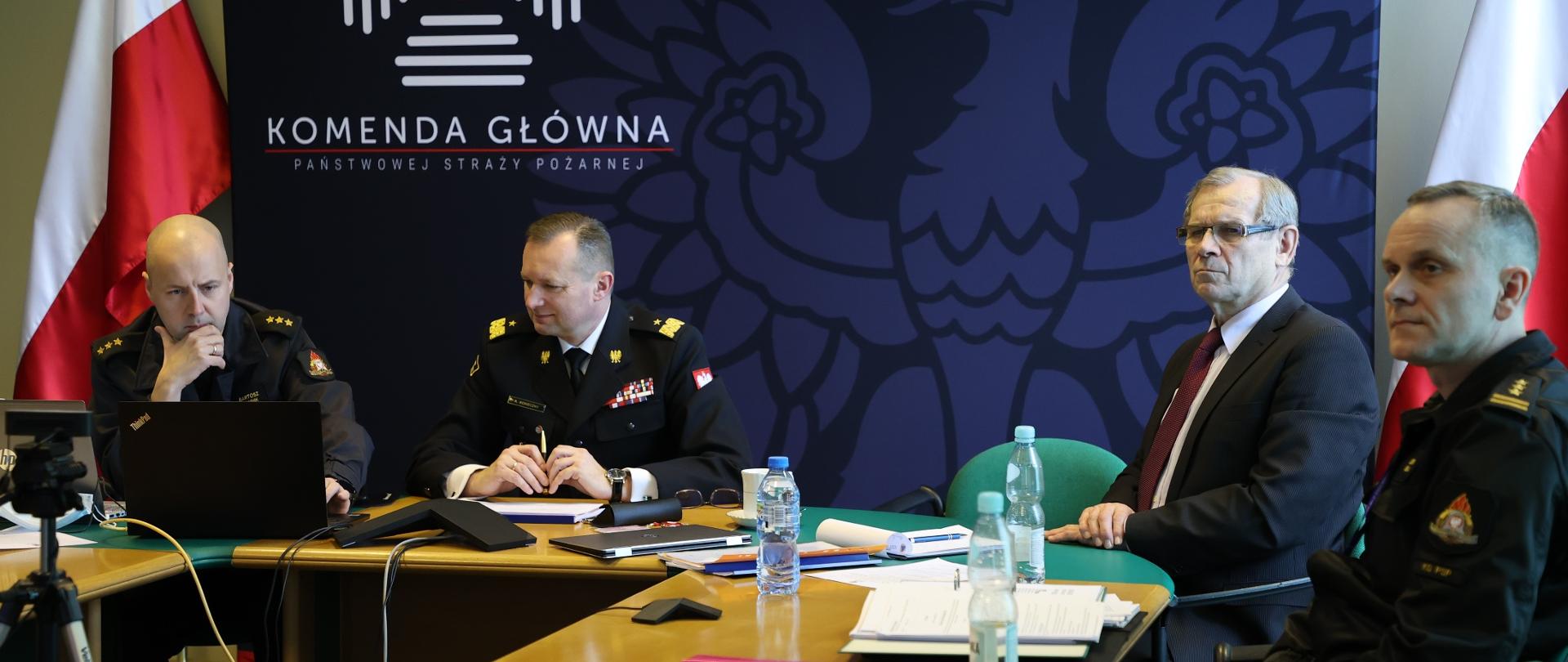 Na zdjęciu widać cztery osoby podczas wideokonferencji. W tle flagi Polski oraz banner z logotypem Komendy Głównej PSP.