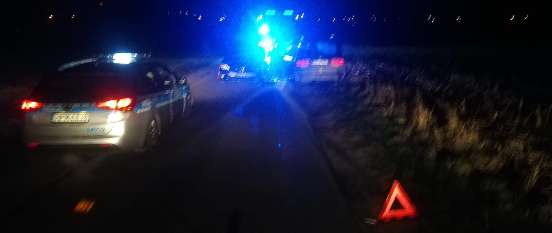 Na zdjęciu widoczne są trzy pojazdy, stojący tyłem na środku drogi radiowóz policyjny oraz stojący tyłem na poboczu drogi samochód osobowy obok którego stoi przodem drugi samochód osobowy. Przed pojazdami na poboczu ustawiony jest trójkąt ostrzegawczy.